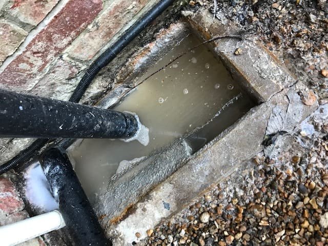 A black water jetting pipe unblocking a flooded rectangular concrete gully drain filled with dirty wastewater next to a exterior brick wall.